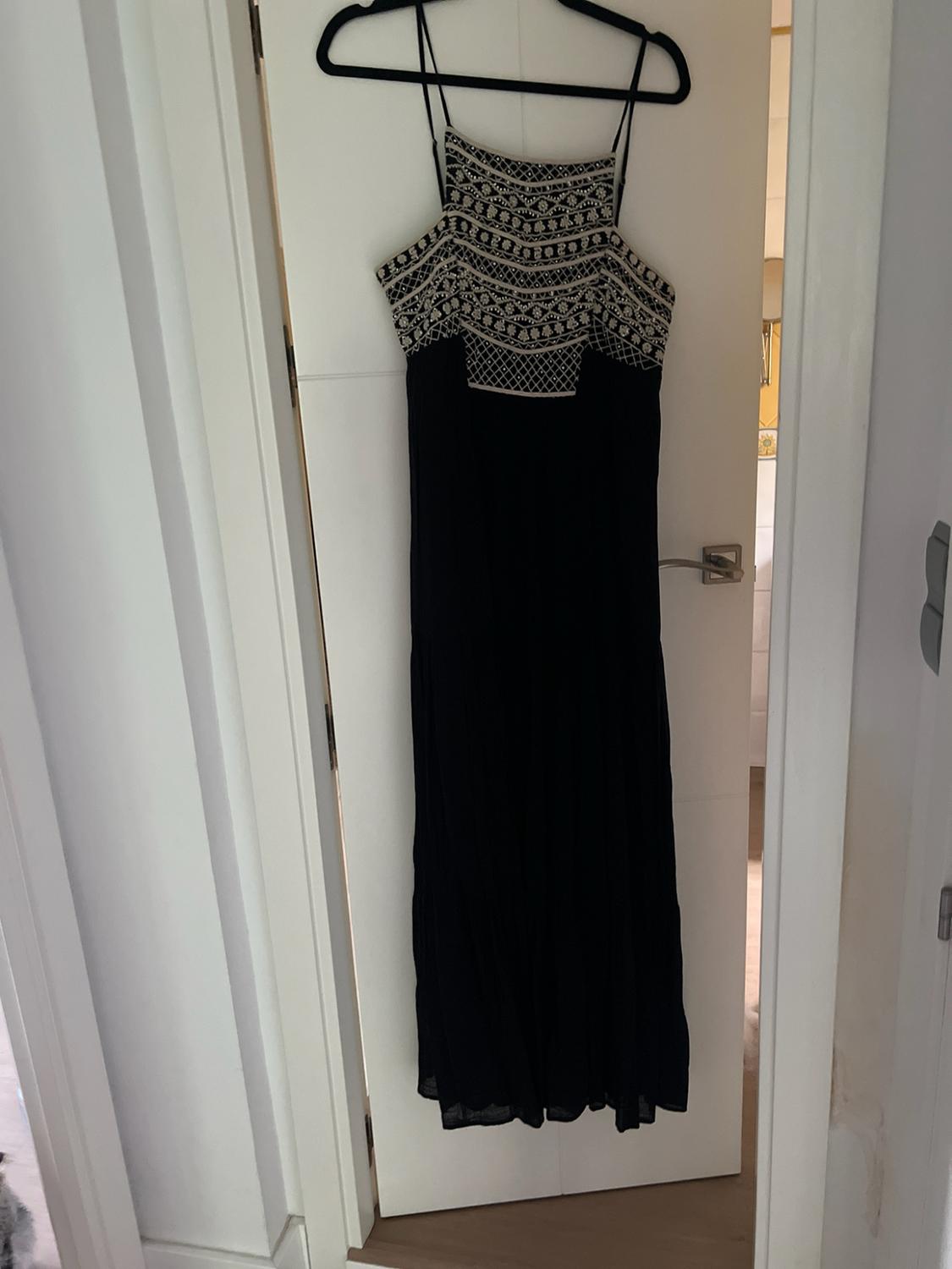 Black maxi dress with straps and geometric embroidery on the chest, hanging on a white door in a relaxed indoor setting.