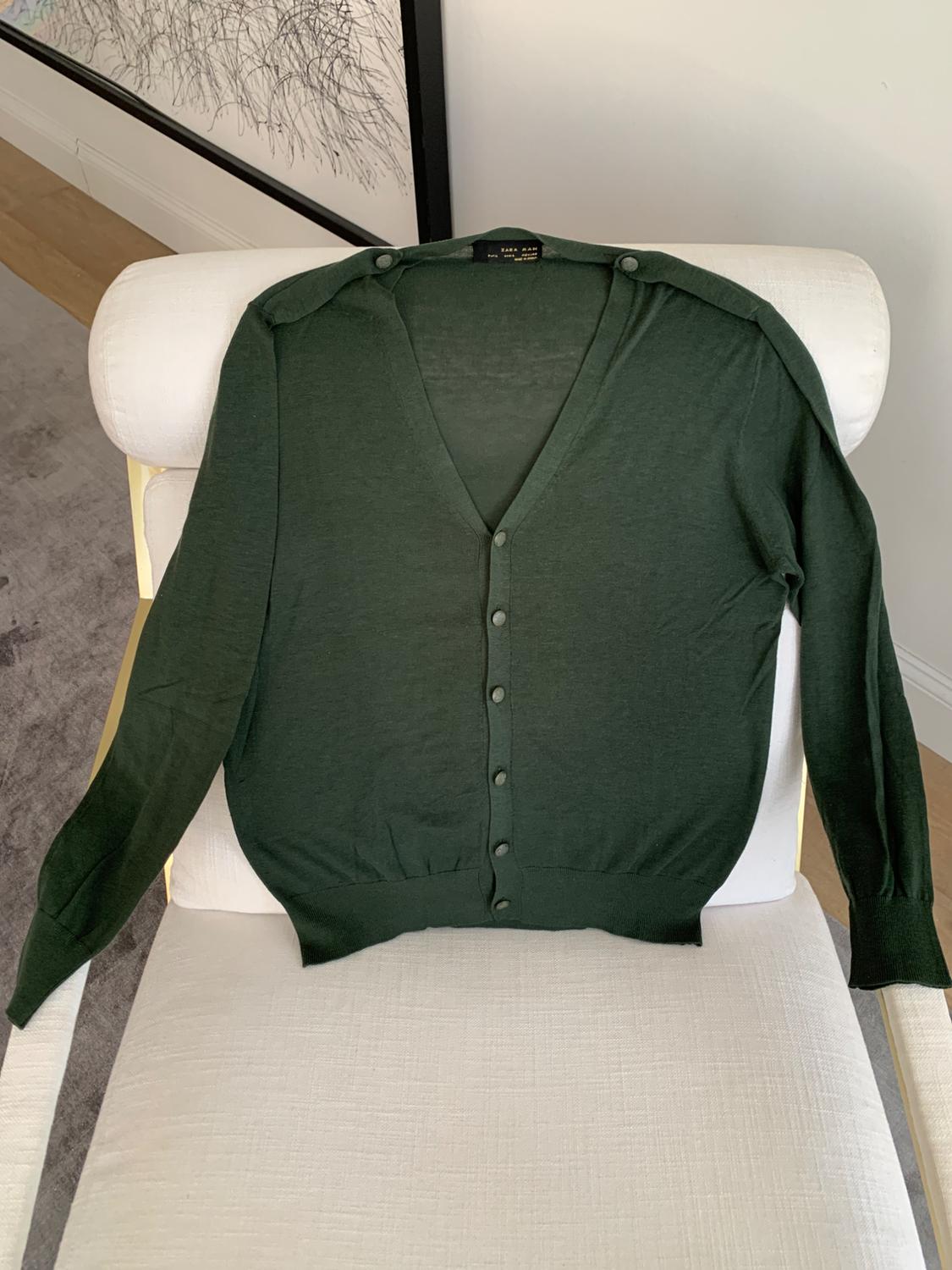 Dark green cardigan with buttons, placed over a white chair. Background with white wall and black and white picture.