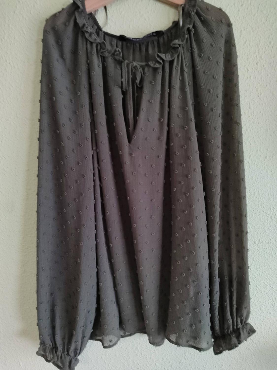 Grey, relaxed-fit blouse with embossed polka dots, ruffled collar, V-neckline and long sleeves, hanging on a hanger against a white wall.
