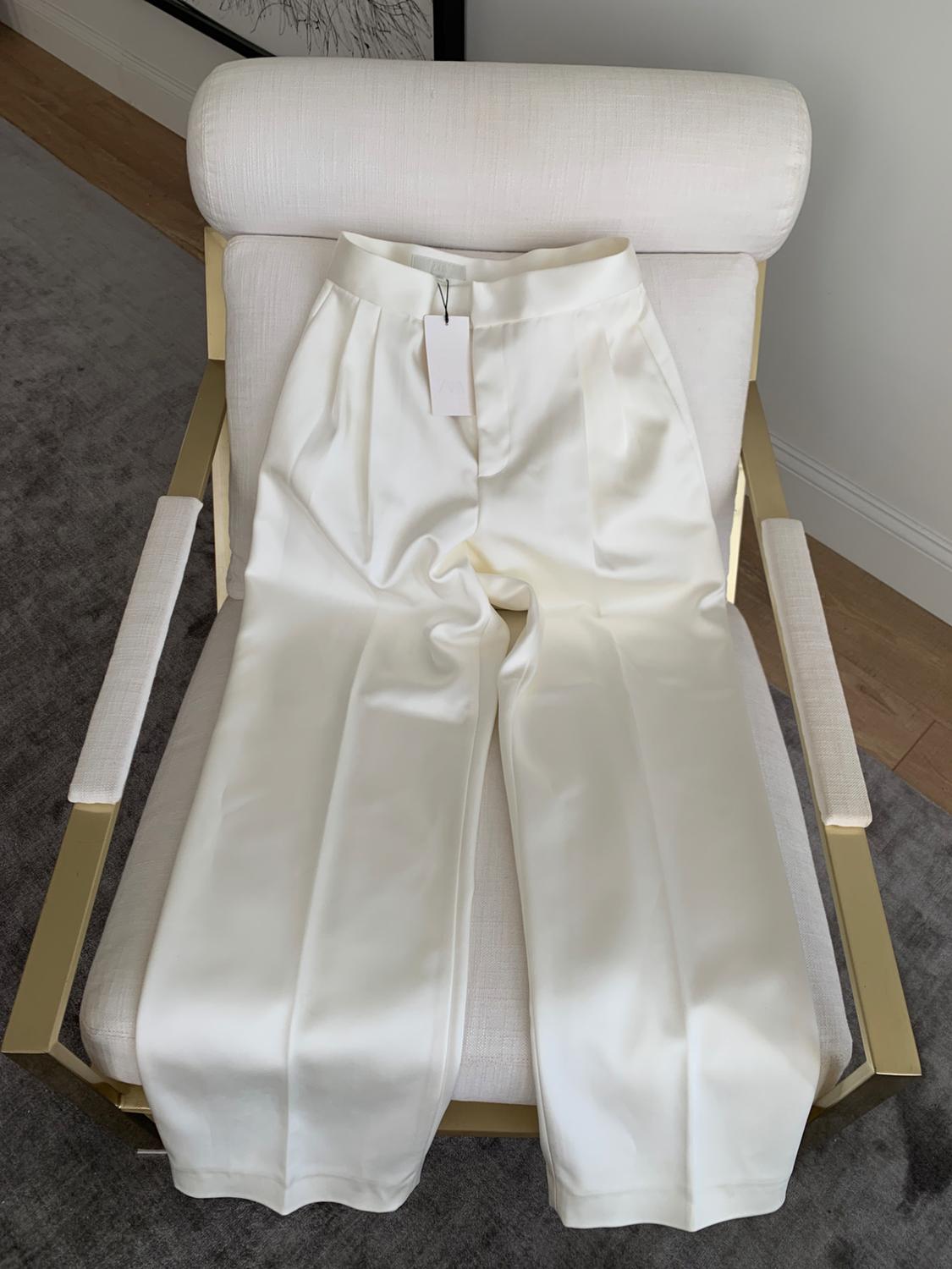 White straight-leg dress pants with a wide leg, with a label, placed on a modern cream-colored chair with gold arms.