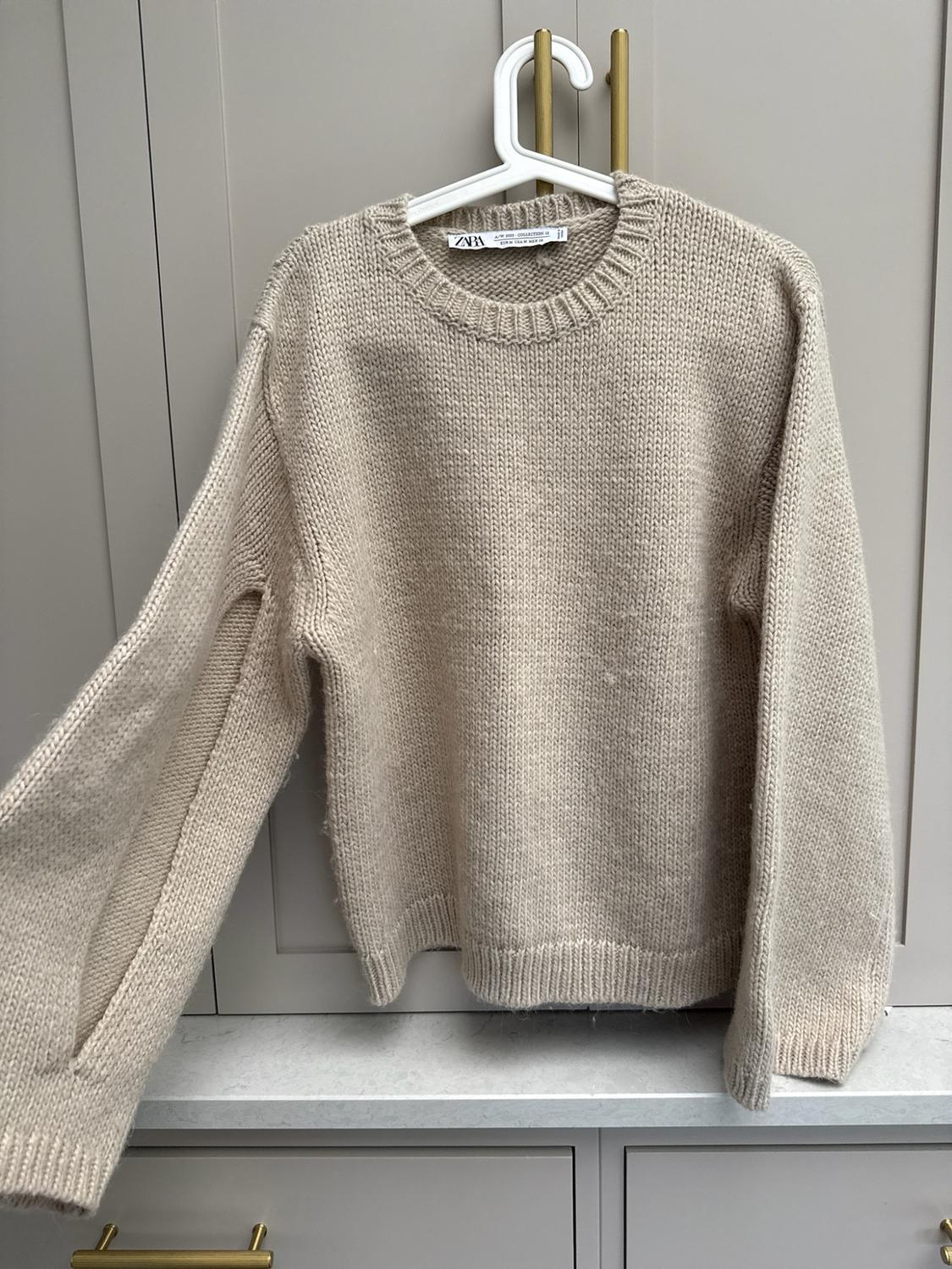 Beige chunky knit jumper hanging on a white hanger over light grey furniture with gold handles. ZARA label visible.