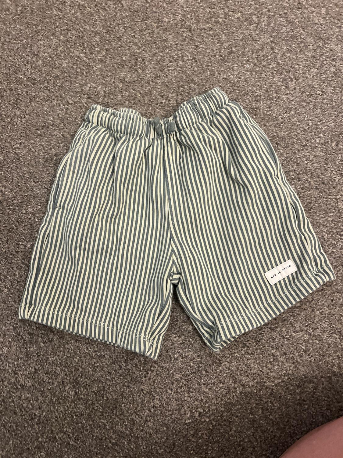 Grey and white striped shorts on a rug. They have an elasticated waist and a patch with the text NYC - LA - TOKYO.