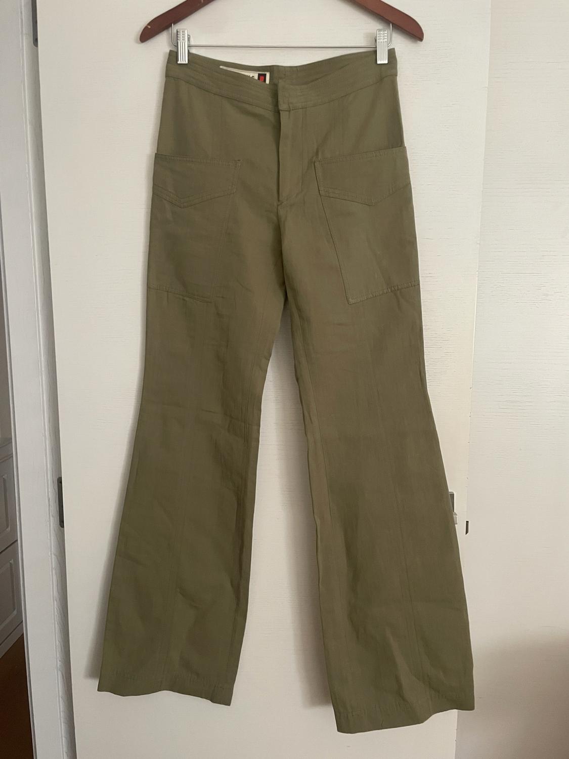 Olive green flared trousers with large front pockets, hanging on a wooden hanger on a white door.
