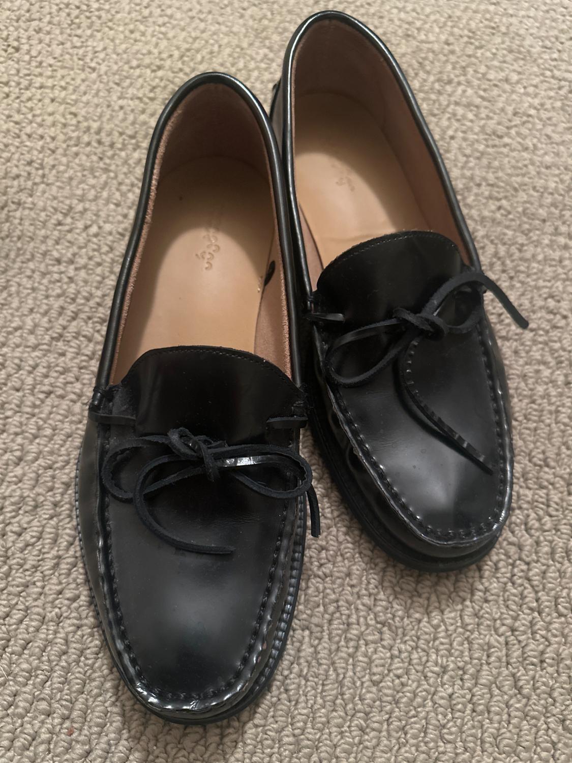 Pair of classic black loafers with a front bow, resting on a textured beige rug. Casual and relaxed style.