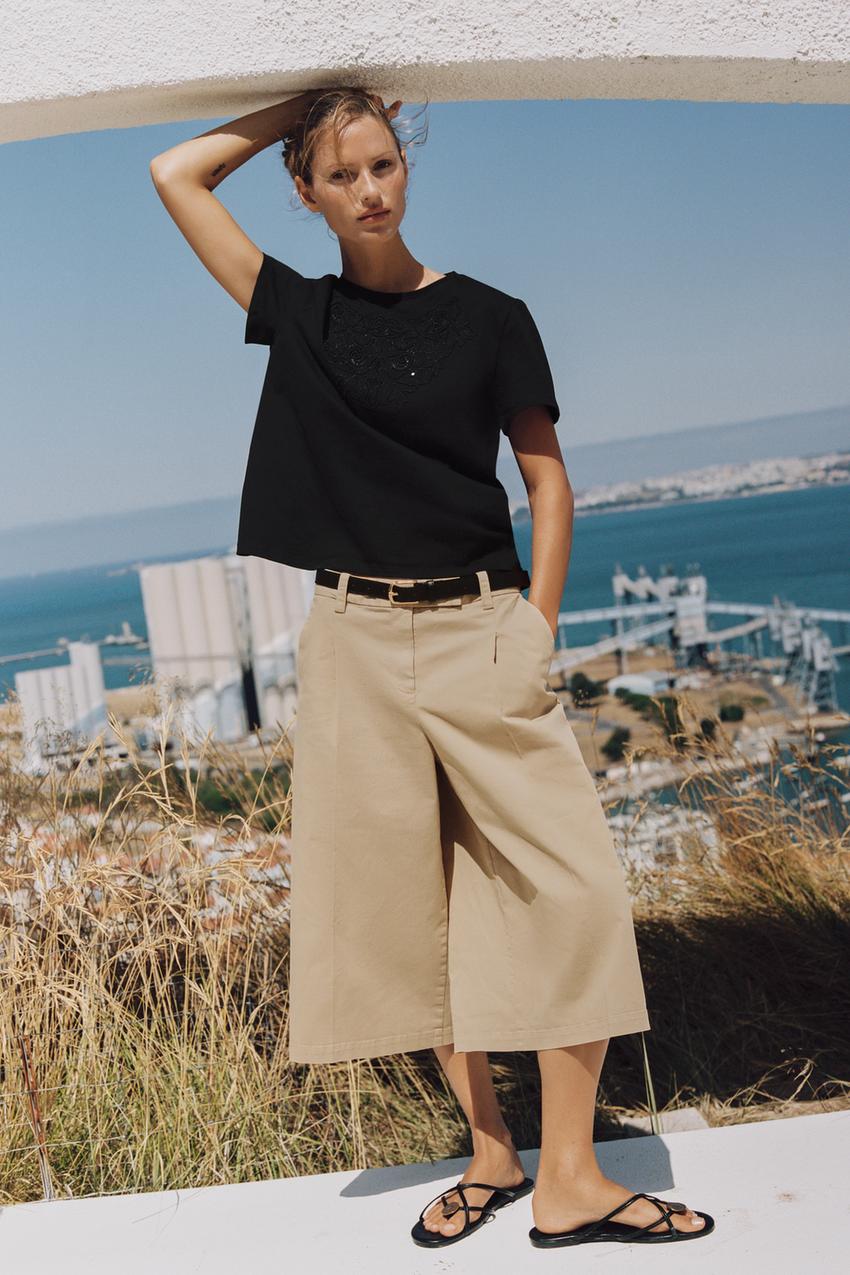 Conjunto de camiseta negra de manga corta con bordado a tono, pantalón beige con bolsillos laterales y cinturón, y sandalias planas.