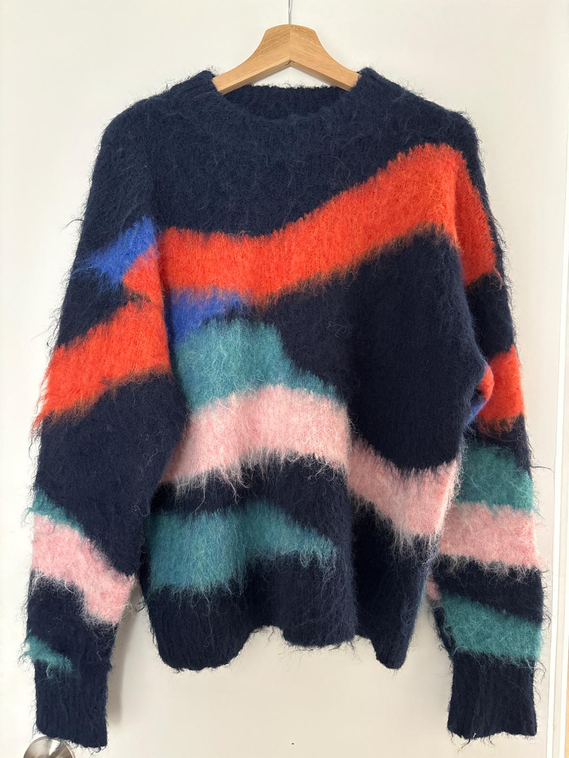 Wool jumper with abstract design in blue, red, pink, and green colours, hanging on a wooden hanger against a white background.