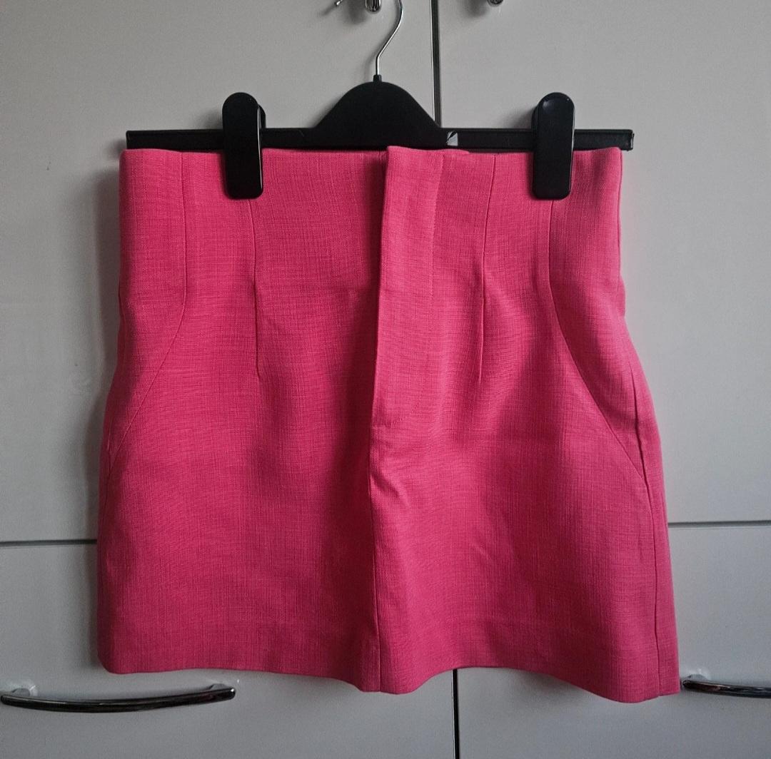 Pink short skirt hanging on a black hanger against a grey wardrobe background. Lightly textured fabric with front pleats.