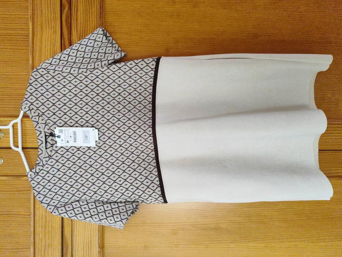 Dress hanging on a white hanger against a wooden background. Top with a geometric pattern and a plain beige skirt. Visible label.