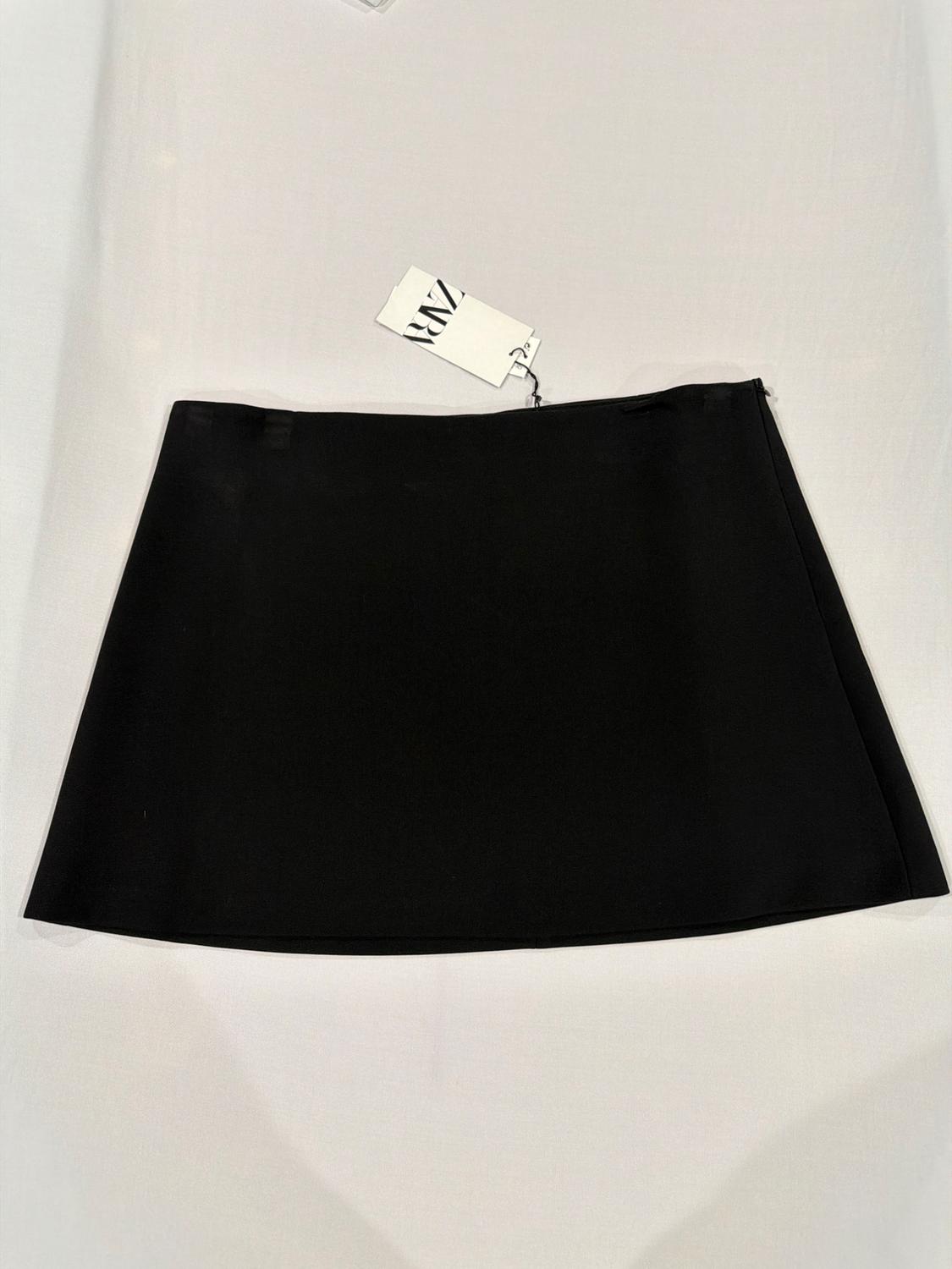 Plain black miniskirt laid out on a white fabric background. A tag with the text ZARA hangs from the waist. Minimalist composition.