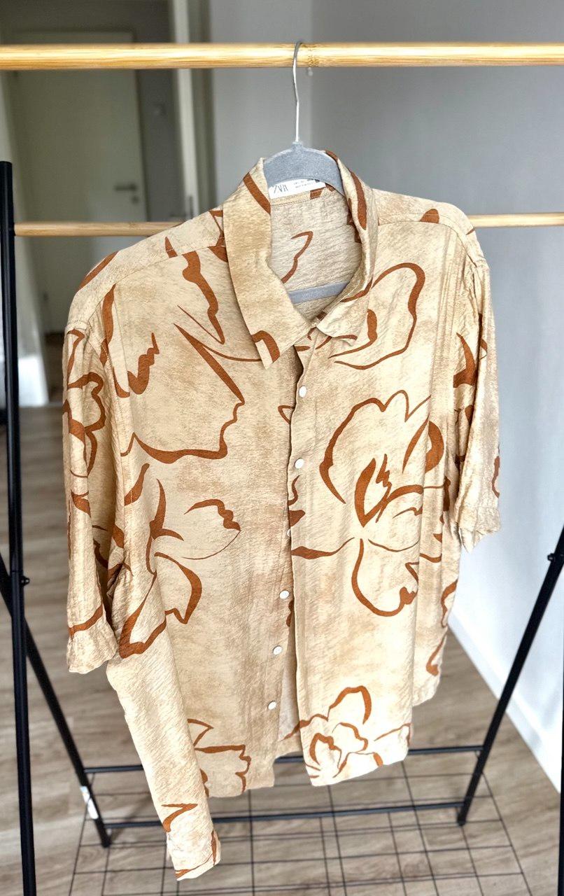 Beige short-sleeved shirt with a brown floral print and ZARA label, hanging on a clothes rack in a room with wooden flooring.
