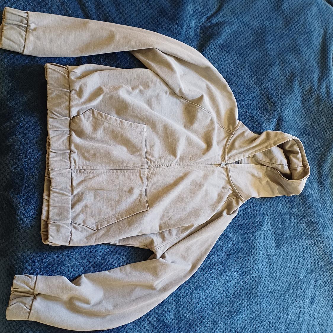 Light beige hooded jacket on a textured blue fabric surface. Long sleeves and front pockets.