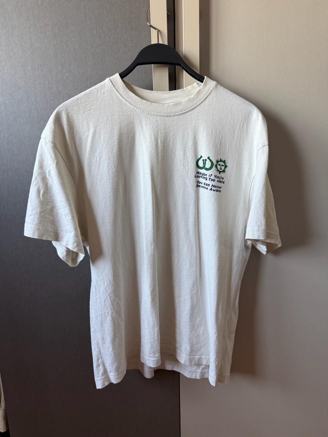 White t-shirt hanging on a hanger. It has a green graphic with the text: "Maybe if You're Looking Too Hard You Can Never Become Aware".
