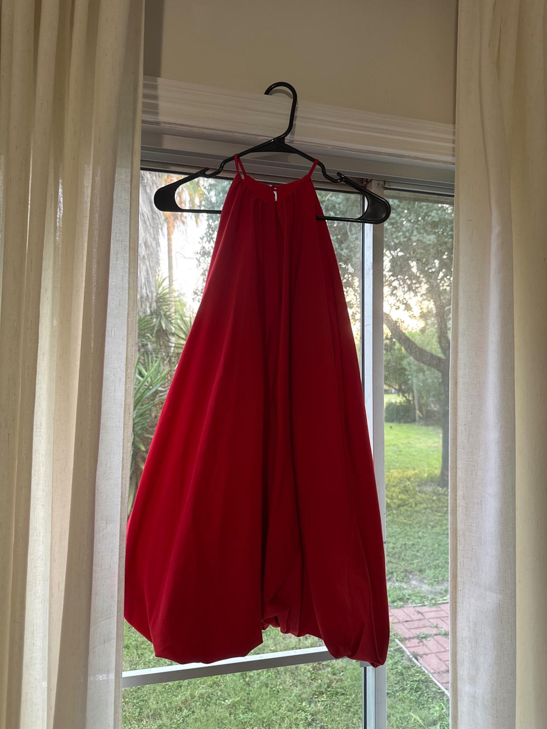 Red dress hanging on a hanger in front of a window, with light curtains and a garden visible in the background.