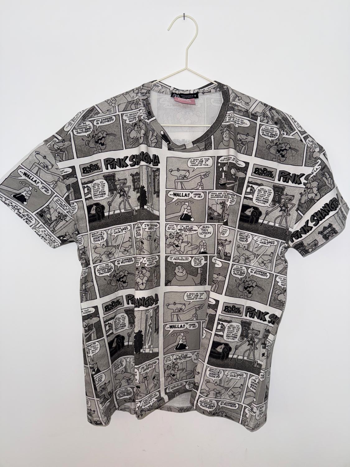 Λευκό κονtomάνικο T-shirt κρεμασμένο σε κρεμάστρα με ασπρόμαυρο τύπωμα κόμικ. Κείμενα: The Pink Panther, PINK SHANGRI-LA.