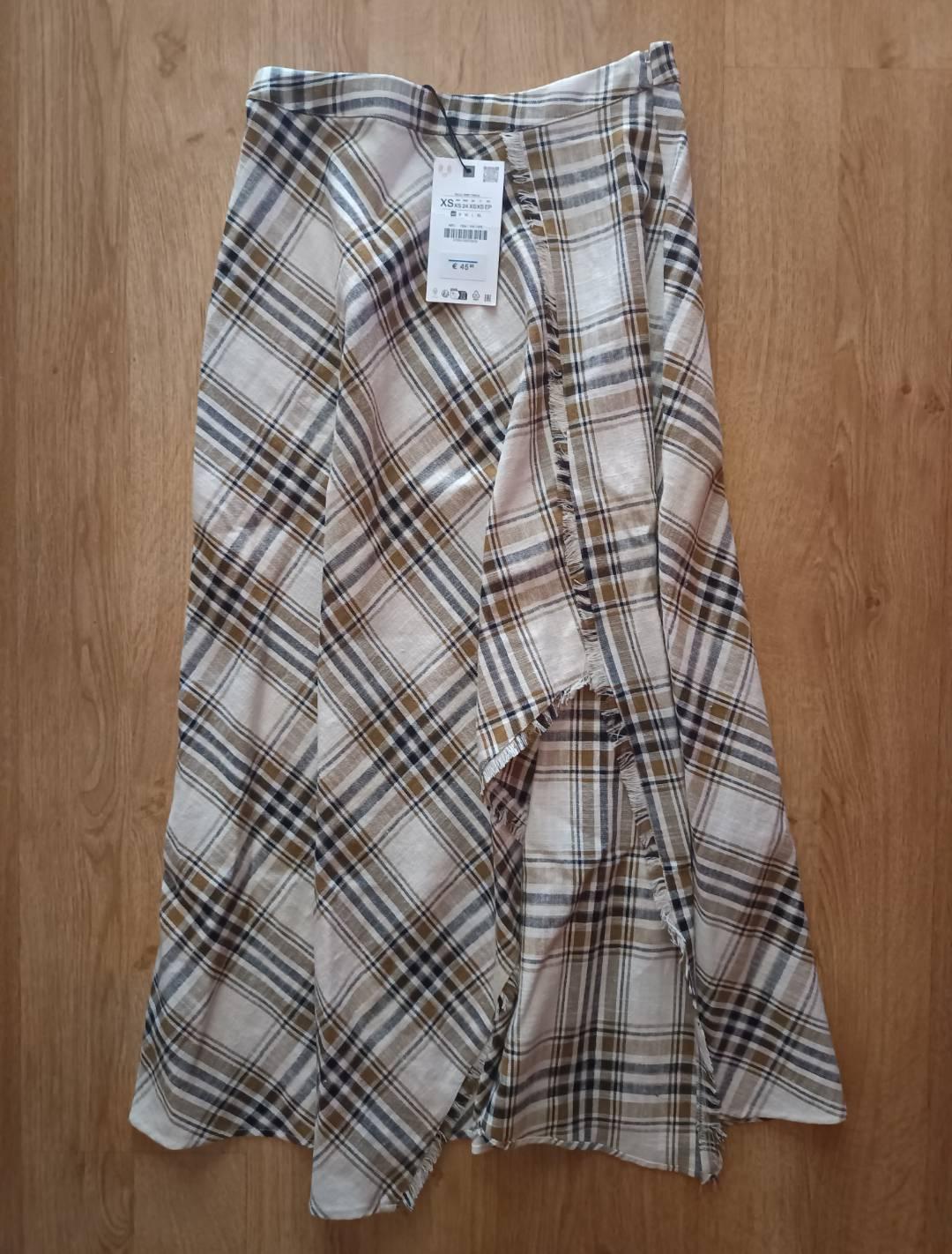 Asymmetrical long checked skirt in white, brown and black with a frayed hem on a wooden floor. Label with text: XS, €45.