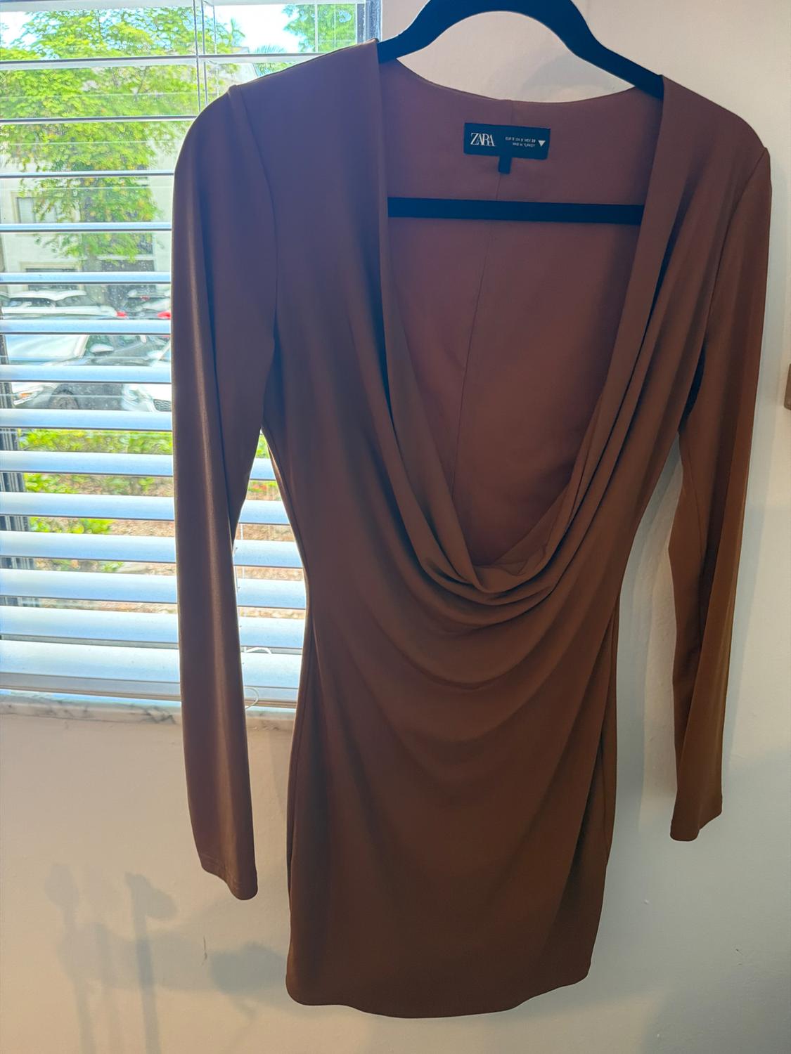 Long-sleeve brown dress with a draped neckline, hanging on a black hanger in front of a window. Tag with the text ZARA.