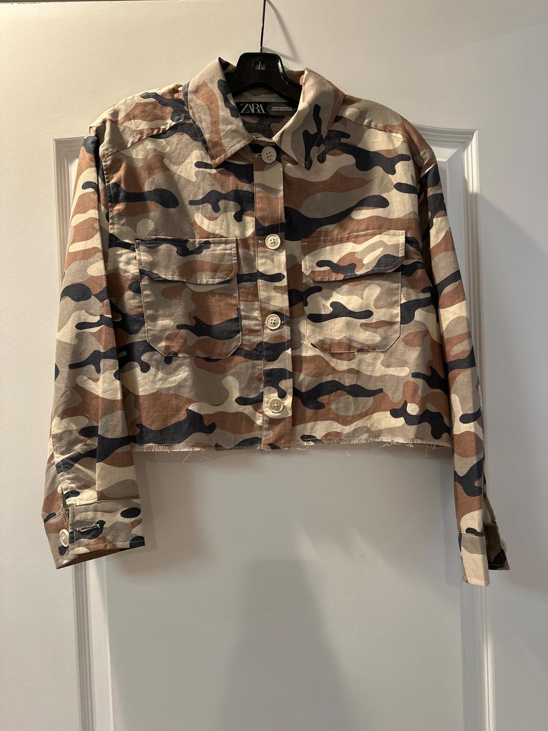 Cropped camouflage jacket in brown and beige tones, with large buttons and two front pockets, hanging on a black hanger.