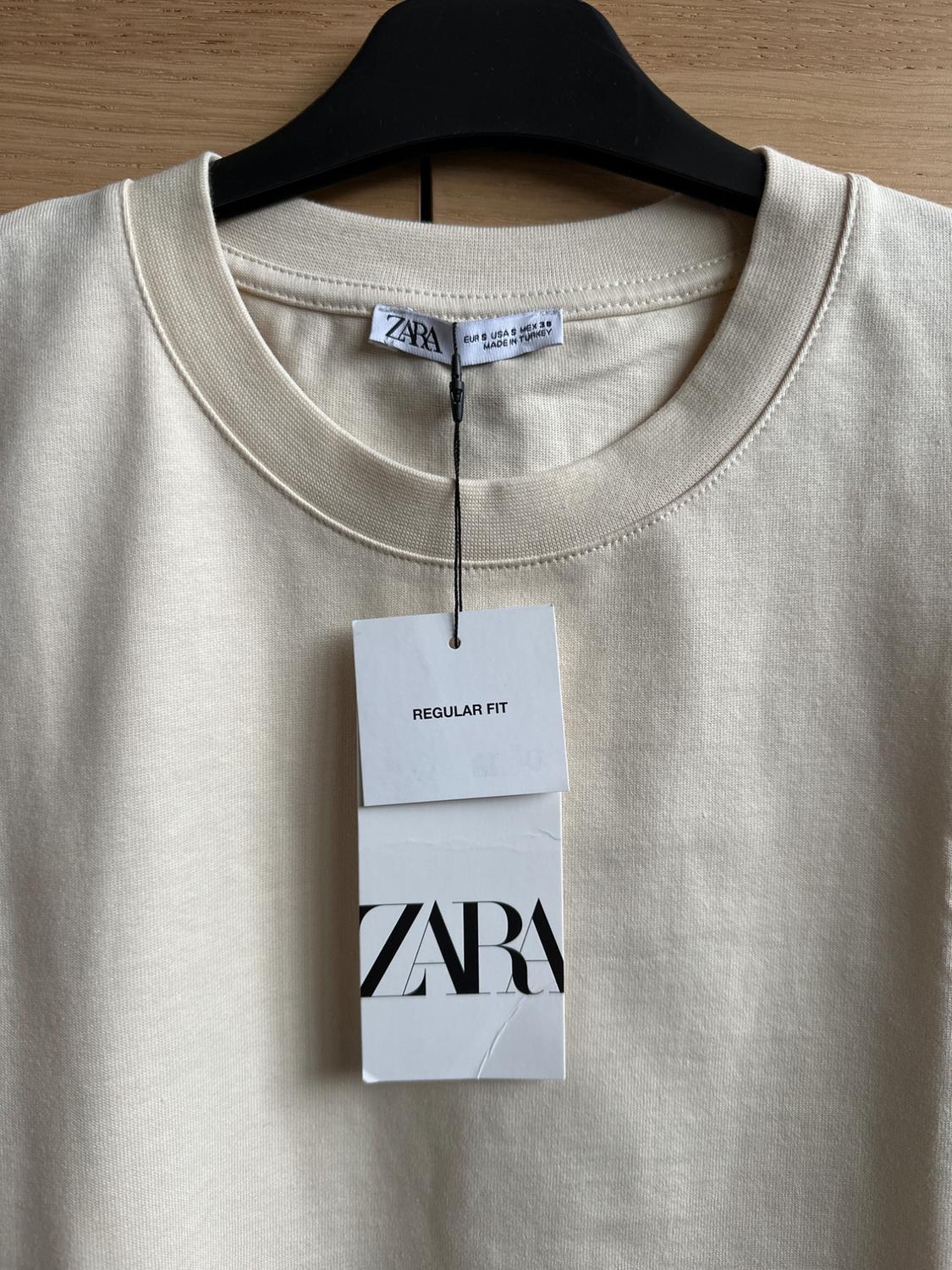 Beige round-neck T-shirt on a black hanger, with a label that says "REGULAR FIT" and "ZARA", against a light wood background.