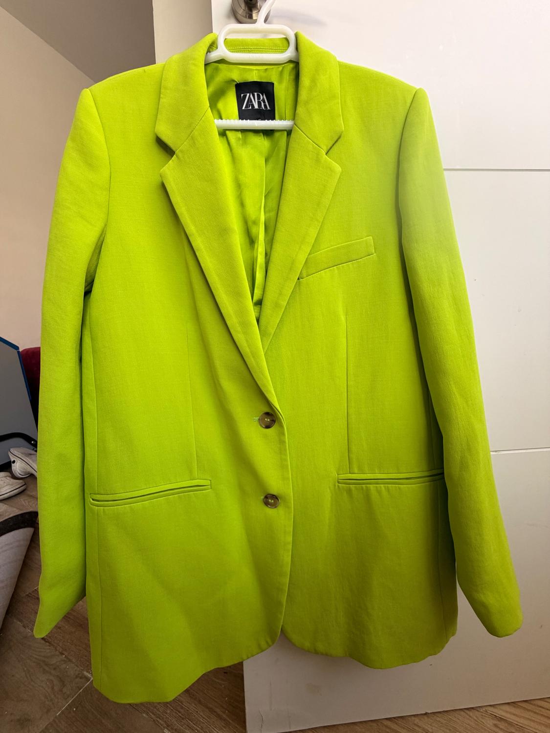 Neon green jacket hanging on a white hanger, with a visible Zara tag, against a white wall background and wooden floor.