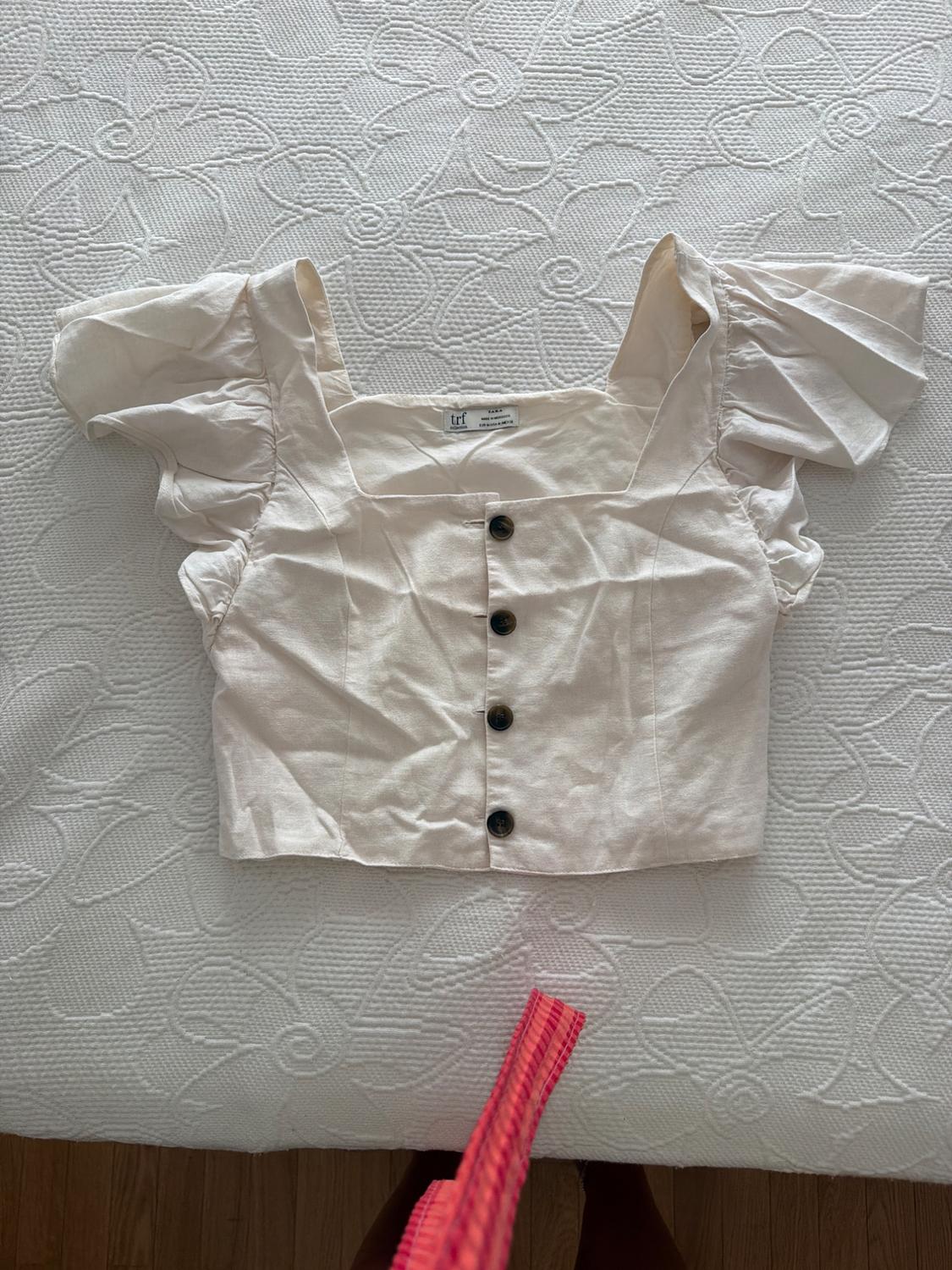 White cropped top with a square neckline, frilled sleeves and front buttons, on a textured white bedspread. Visible text: trf ZARA.