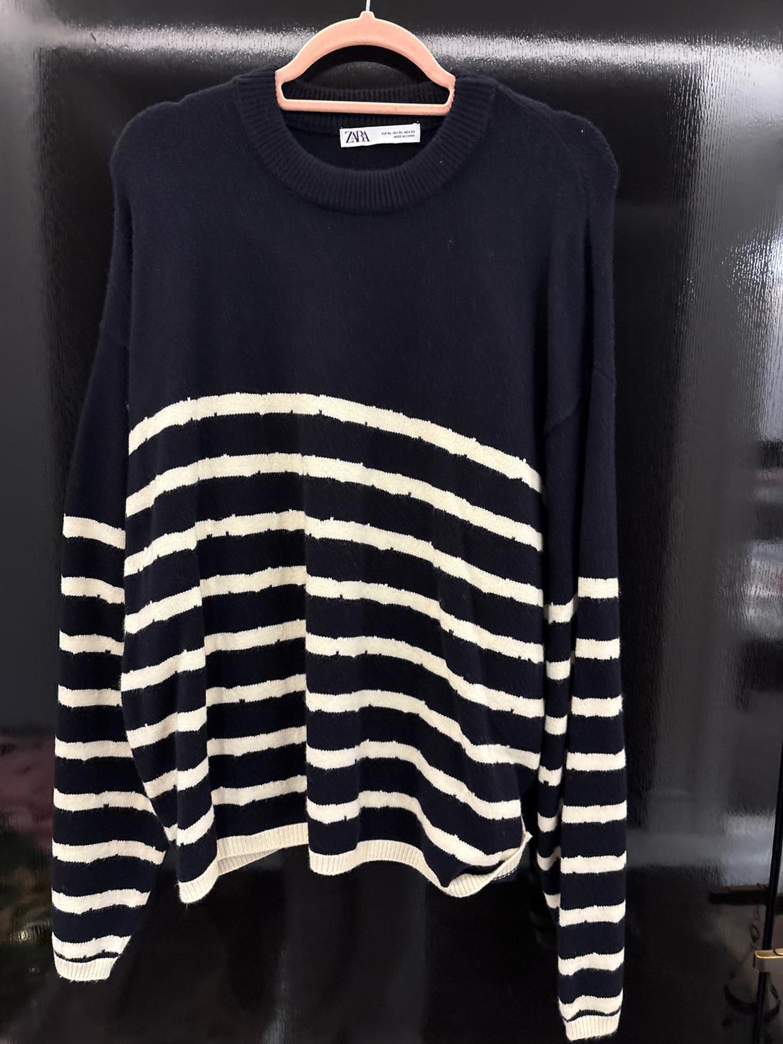 Black and white striped jumper hanging on a wooden hanger. Visible label with the text "ZARA". Shiny black background.