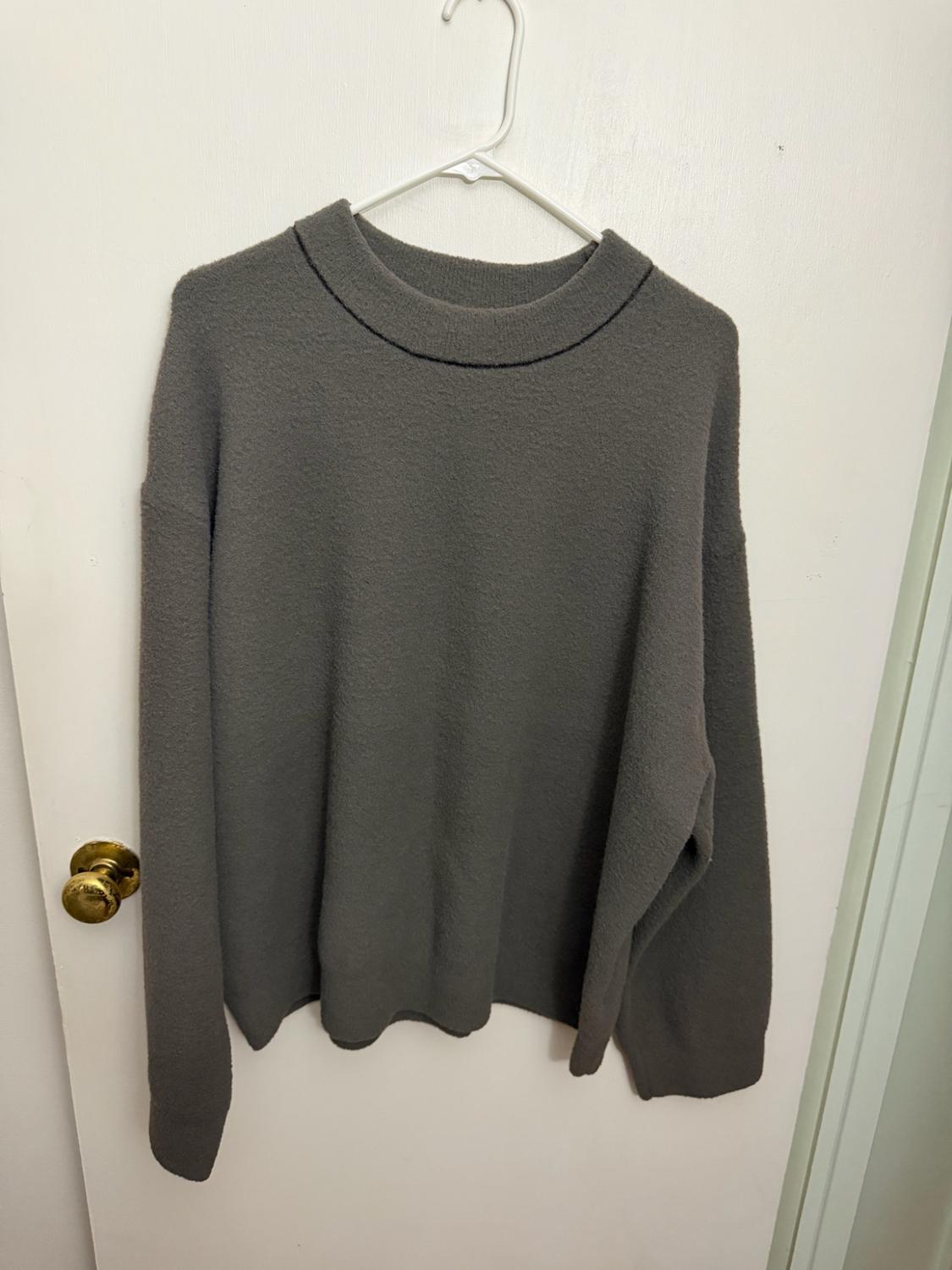 Dark gray long-sleeved knit sweater hanging on a hanger on a white door with a gold knob. Casual and relaxed style.