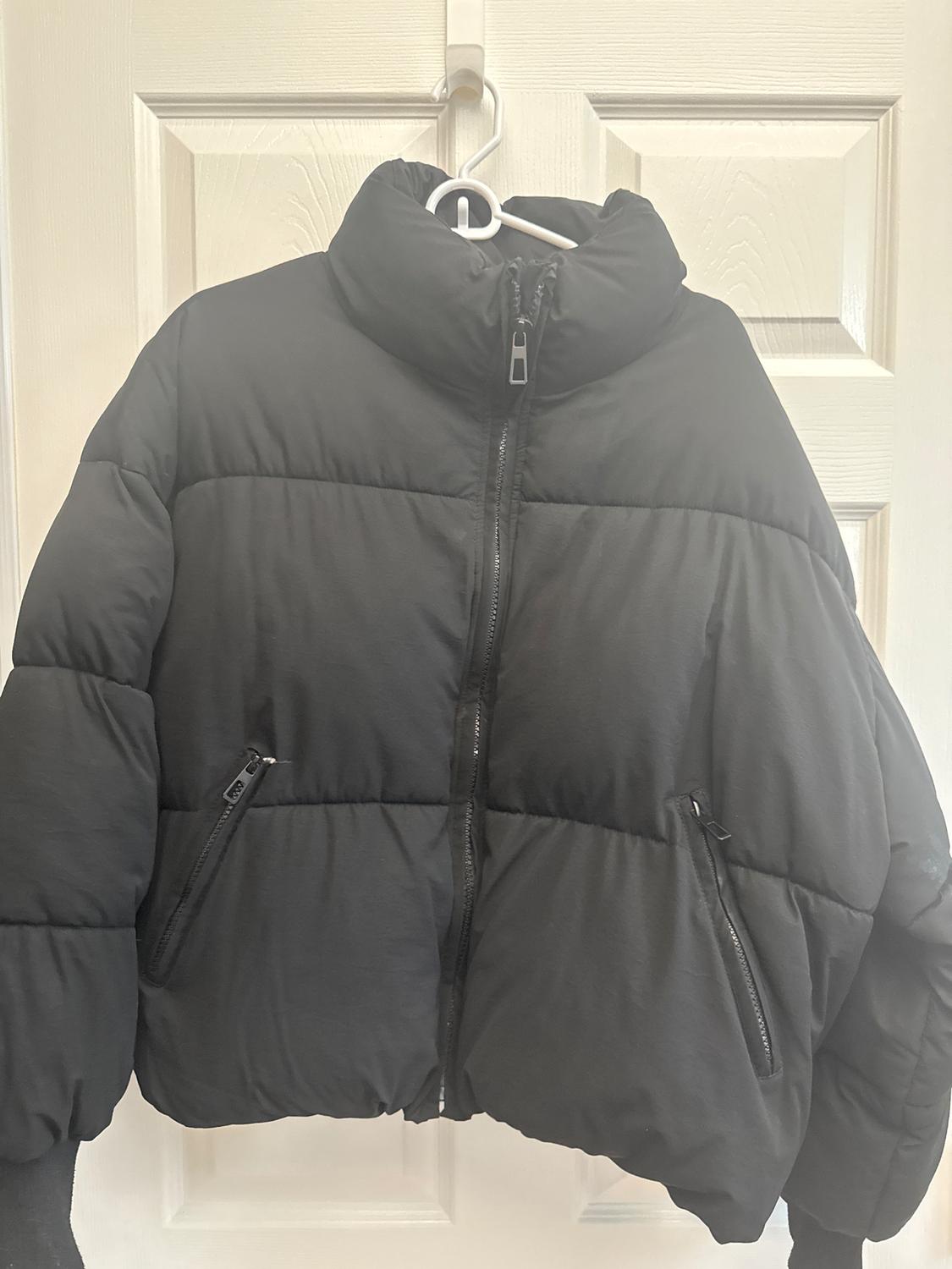 Black quilted jacket hanging on a white hanger over a white door. It has a central zip and side pockets.