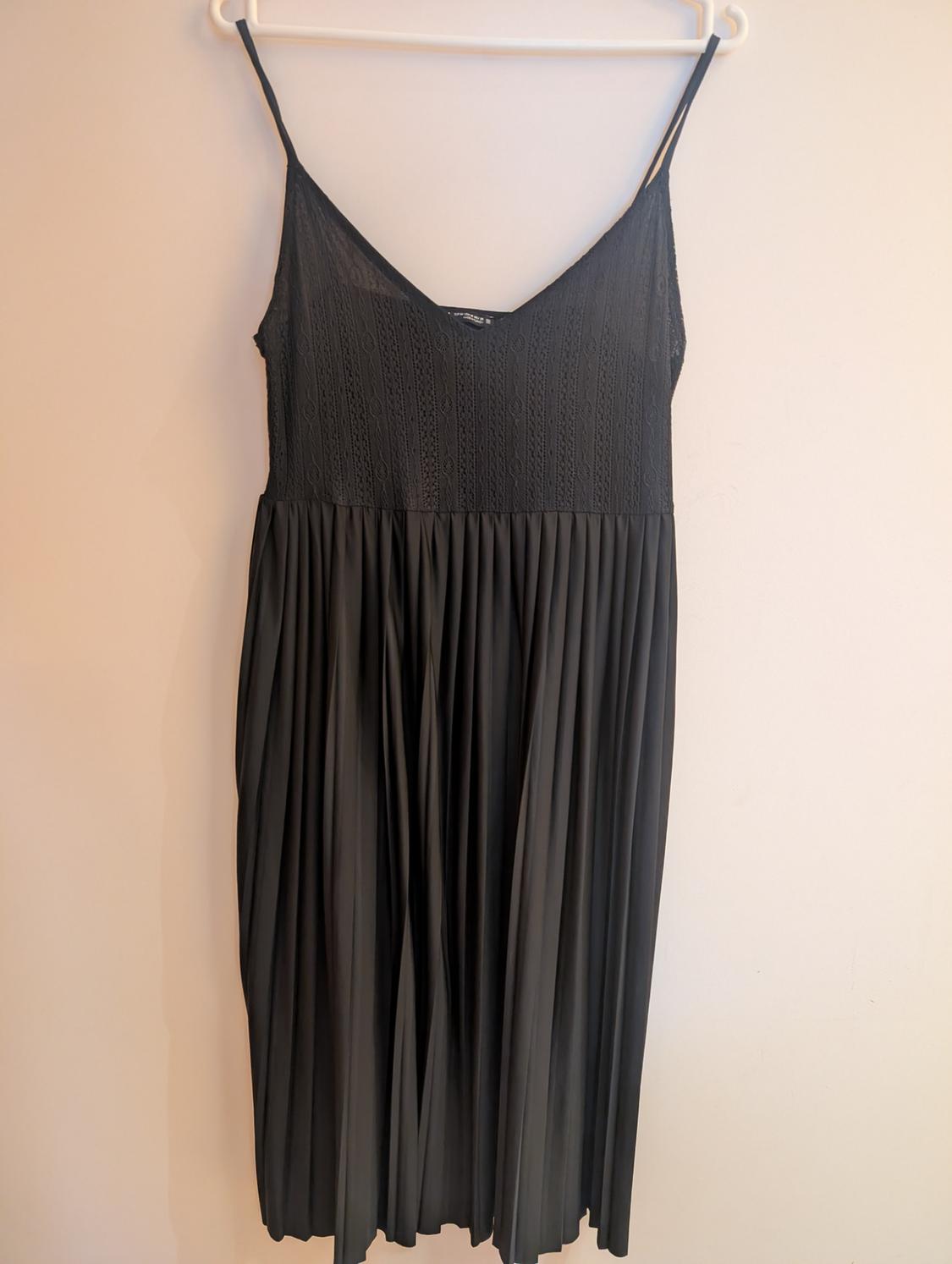 Black dress with spaghetti straps, V-neck, textured bodice and pleated skirt. It is hanging on a white hanger against a light-coloured wall.