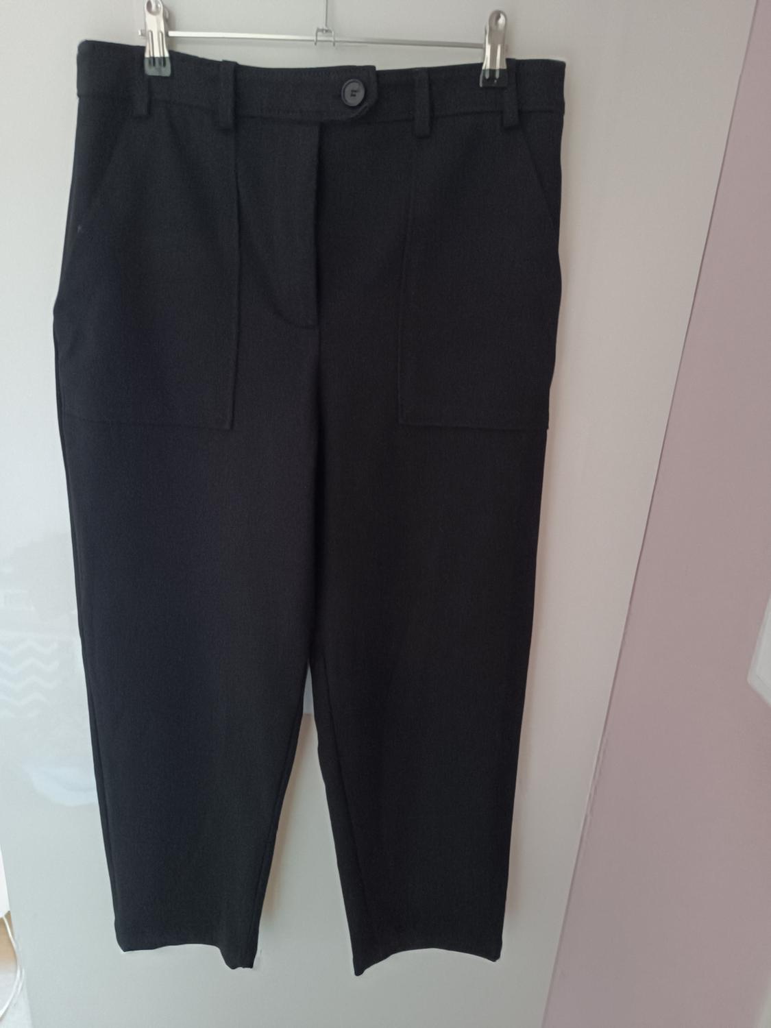 Black trousers hanging on a hanger against a light background. Simple design with front pockets and button fastening.