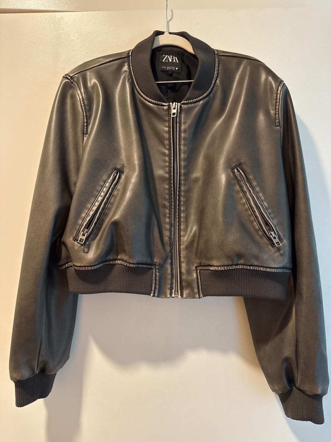 Black faux leather jacket hung on a hanger, with front closure and zippered pockets. Visible tag: ZARA.