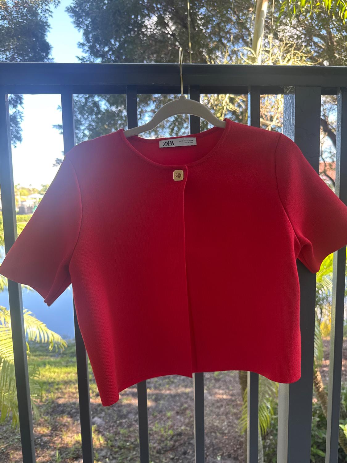 Red short-sleeve jacket hanging on a hanger in front of a railing, with a background of trees and blue sky. Visible tag.