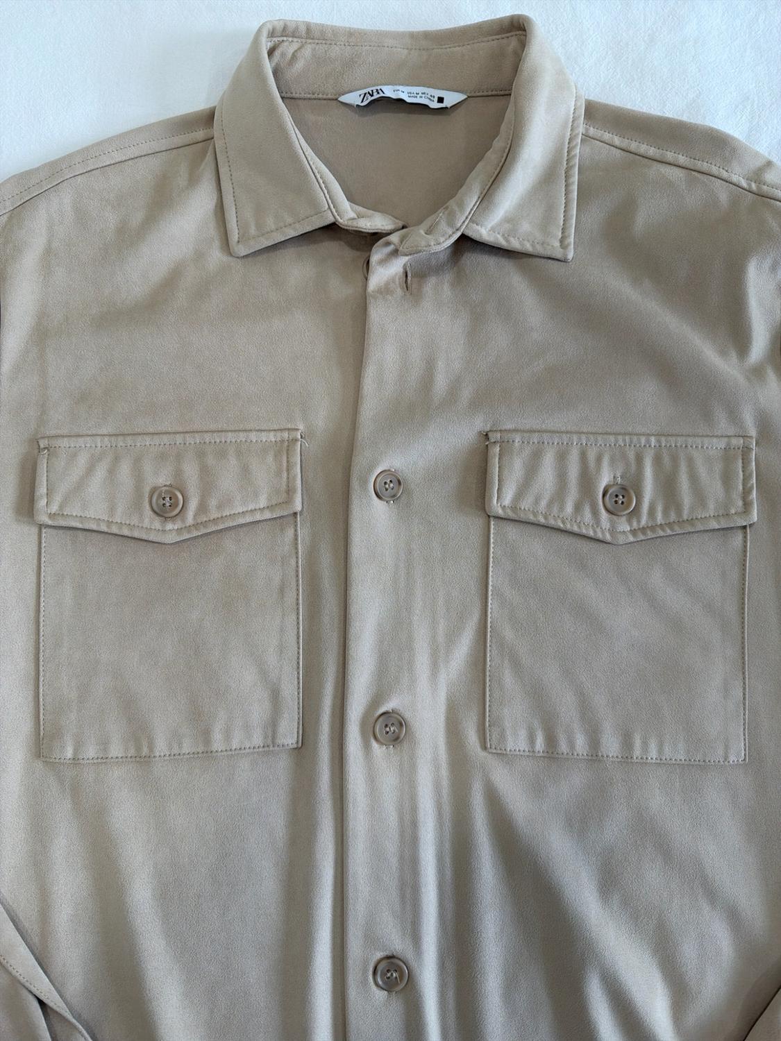 Close-up of beige shirt with a collar, button front, and two flap pockets on the chest. Inner label with ZARA text.