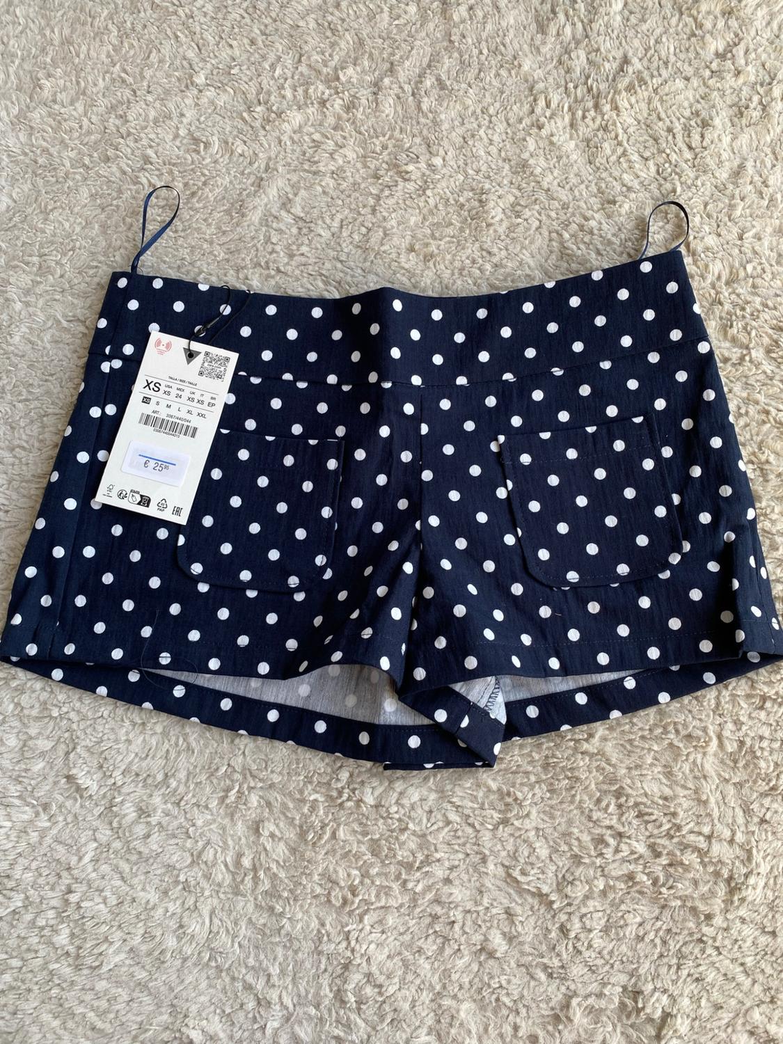 Navy blue shorts with white polka dots on a carpet. They have back pockets and a label displaying the text XS and €25.95.