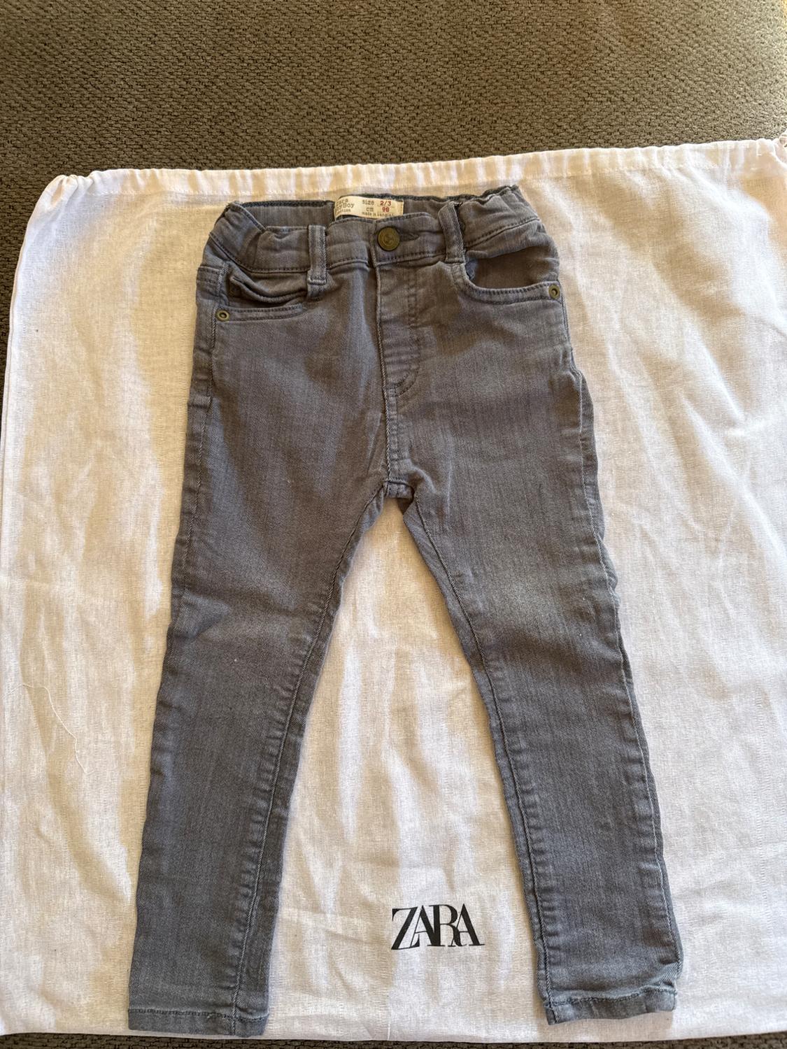 Grey children's trousers on a white bag with the "ZARA" logo at the bottom. Textured fabric background.