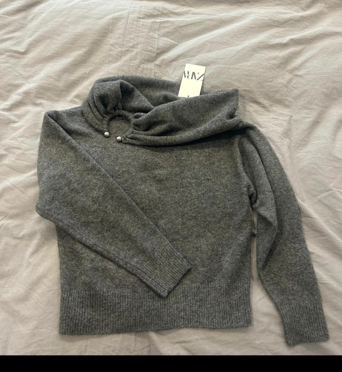 Grey long-sleeved jumper with adjustable collar and drawstring detail, on a grey surface. Visible label.