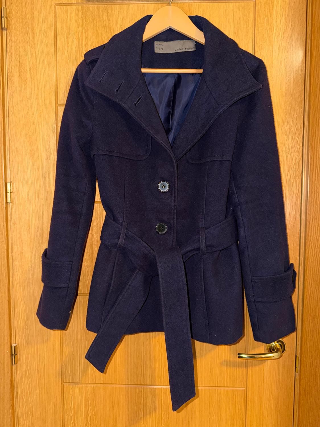 Dark blue coat with a belt and buttons, hanging on a wooden hanger over a light wooden door. Visible label: ZARA BASIC.
