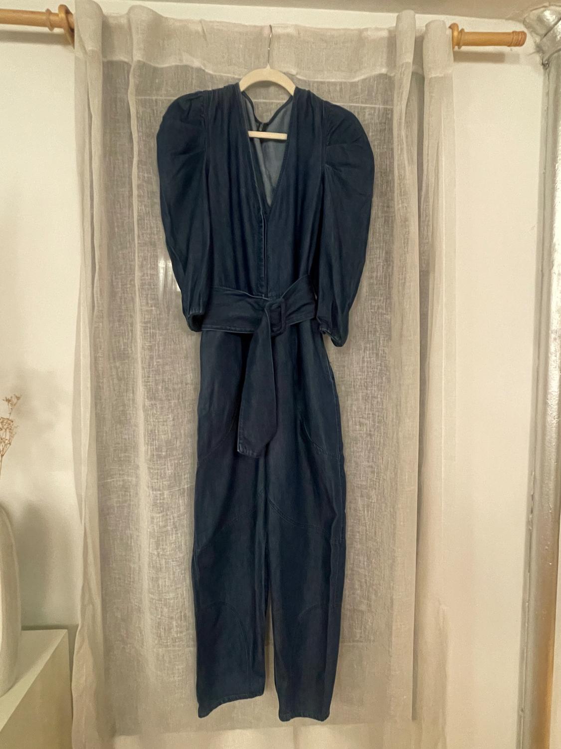 Dark blue jumpsuit with puffed sleeves and a belt, hanging on a hanger in front of a beige translucent curtain.