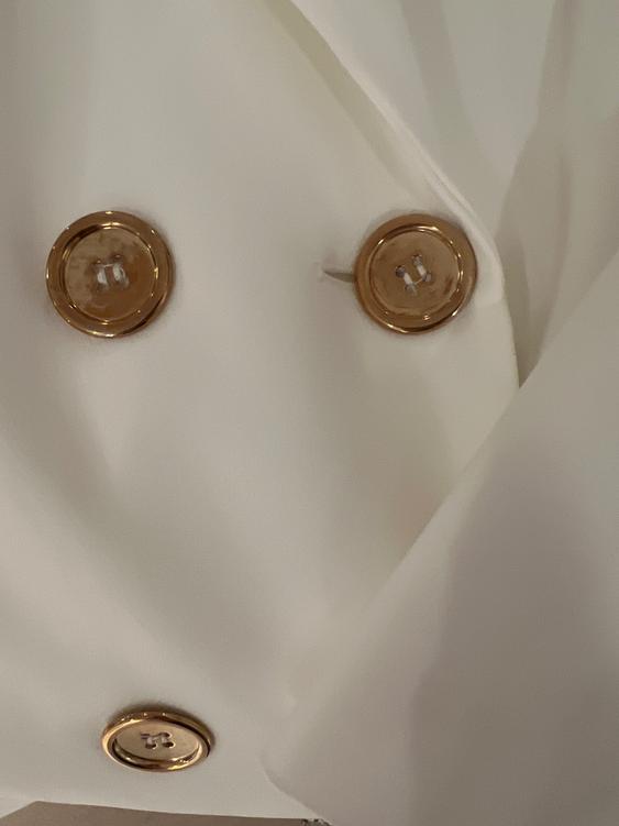 Detail of a white coat with shiny gold buttons. The fabric has a soft and elegant finish.