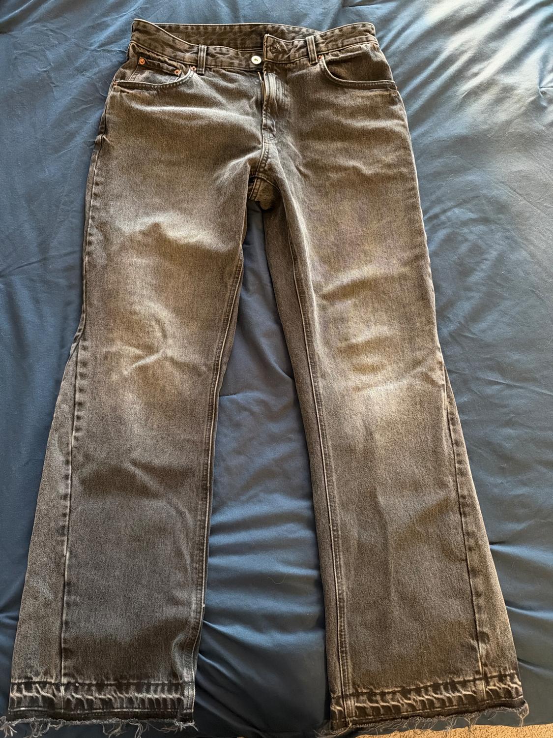 Dark gray denim jeans with a washed effect, a straight cut, and frayed hem, spread out over a blue surface.