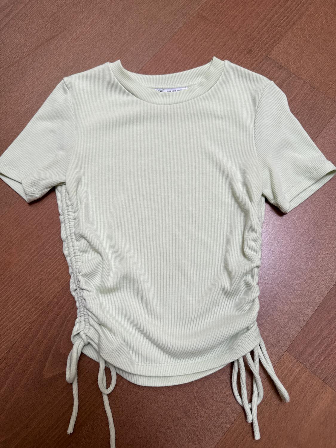 Beige short-sleeved T-shirt with side ruching and drawstrings, on a brown wooden floor.