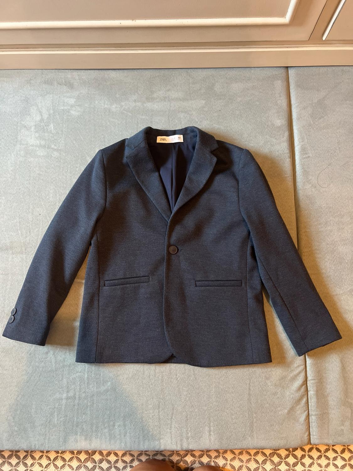 Dark blue jacket with Zara text on the label, on a grey surface. Above there is a white piece of furniture and below there is a patterned rug.