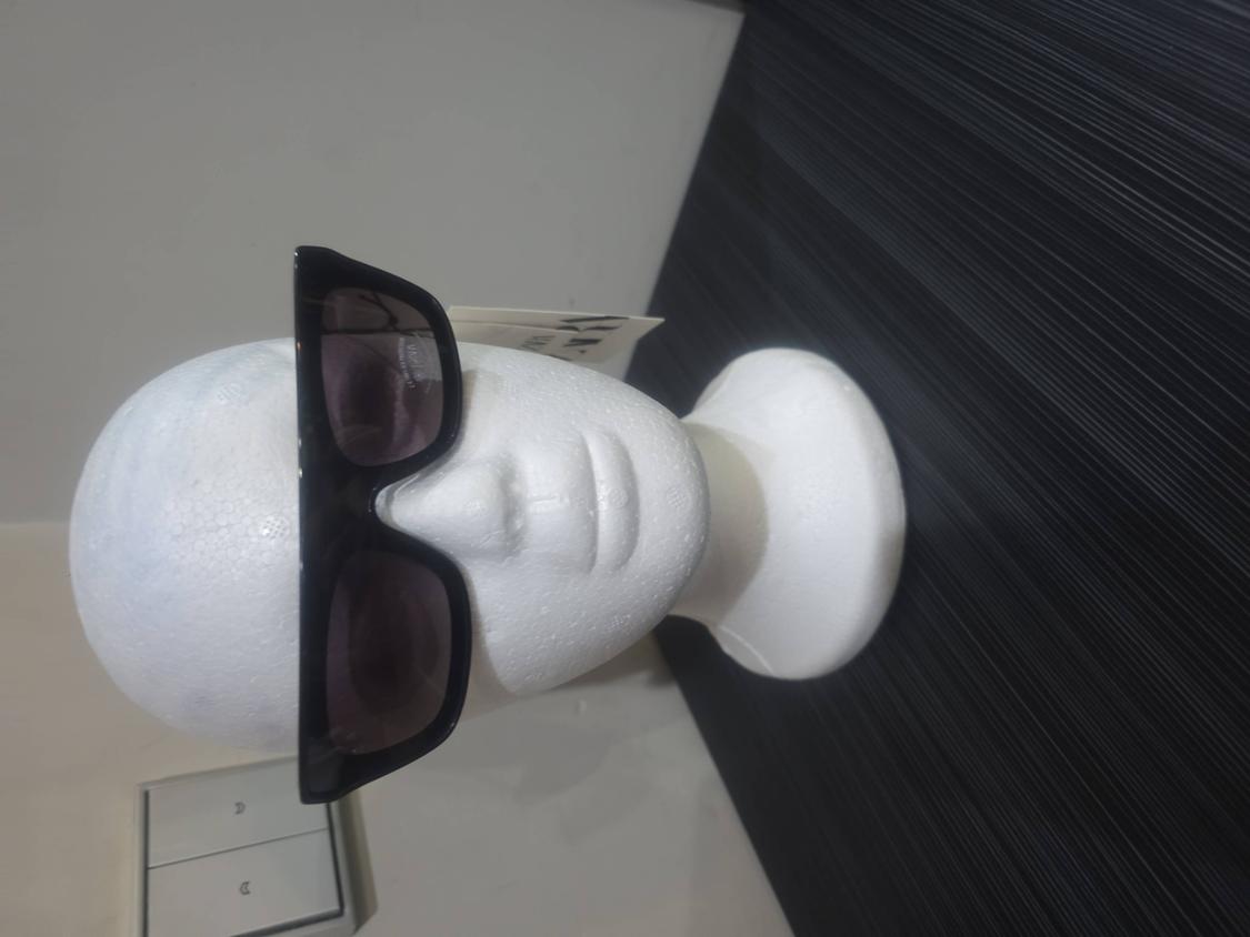 Black rectangular sunglasses on a white mannequin head. Background split between a light wall and a dark textured panel.