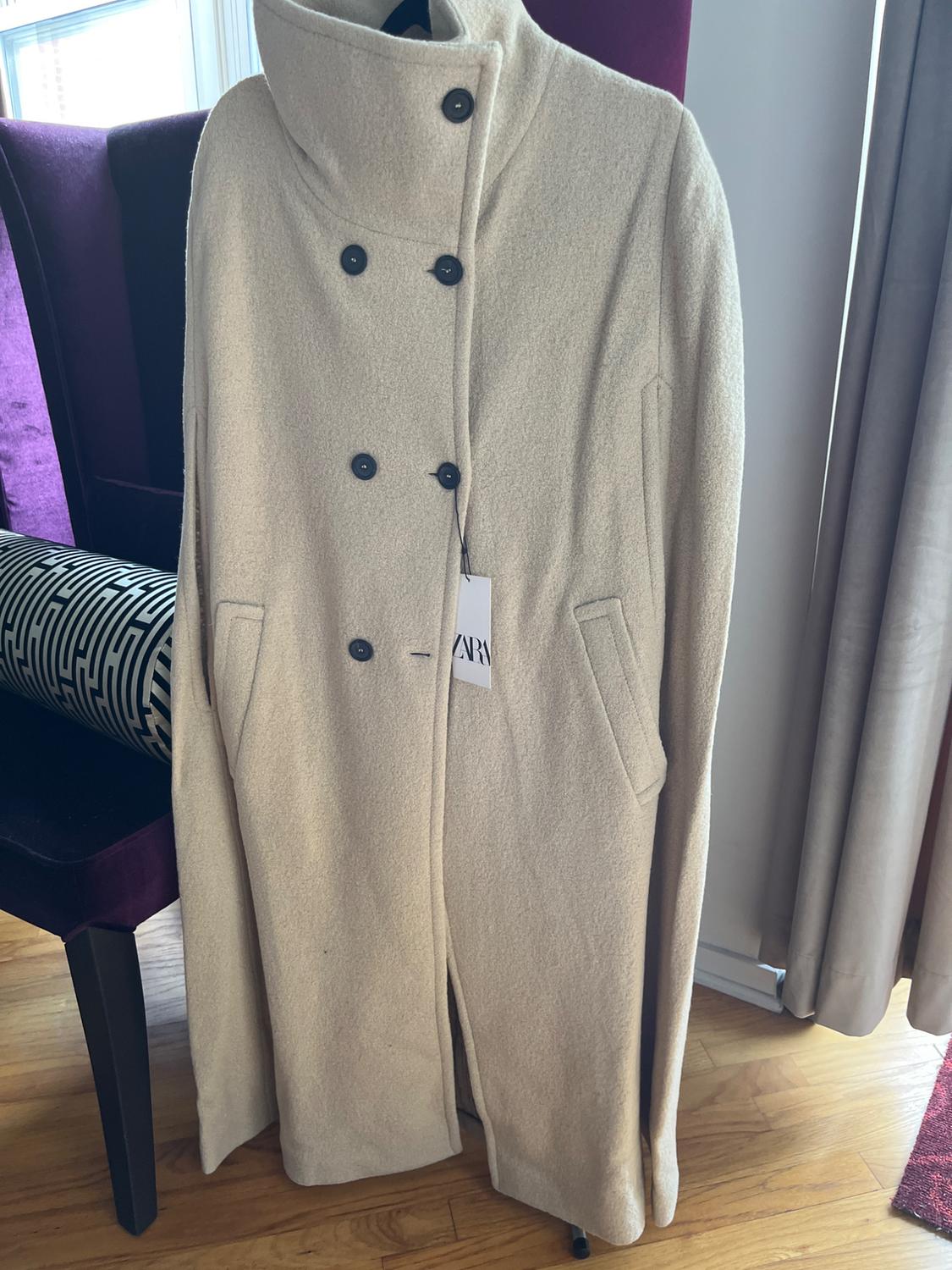 Beige coat with a high collar and black buttons, hanging on a purple chair. Visible label with "ZARA" text. Background with curtains and wooden floor.