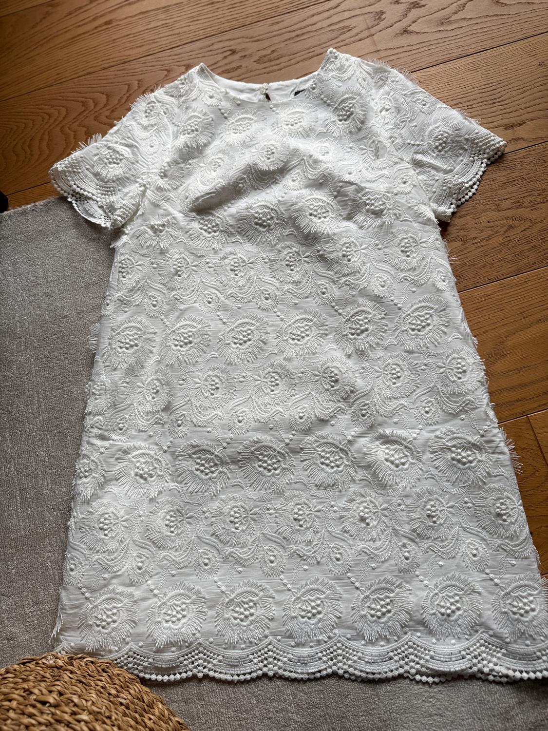 White lace dress with a floral design on a wooden background and a grey rug. Scalloped edge detail.