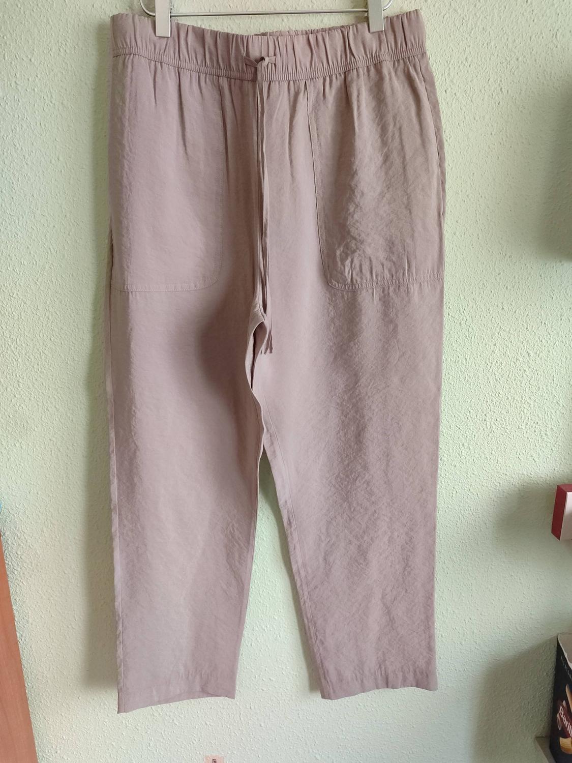 Dusty pink trousers with an elasticated waist, drawstring and front patch pockets, hanging on a metal hanger against a light-coloured wall.