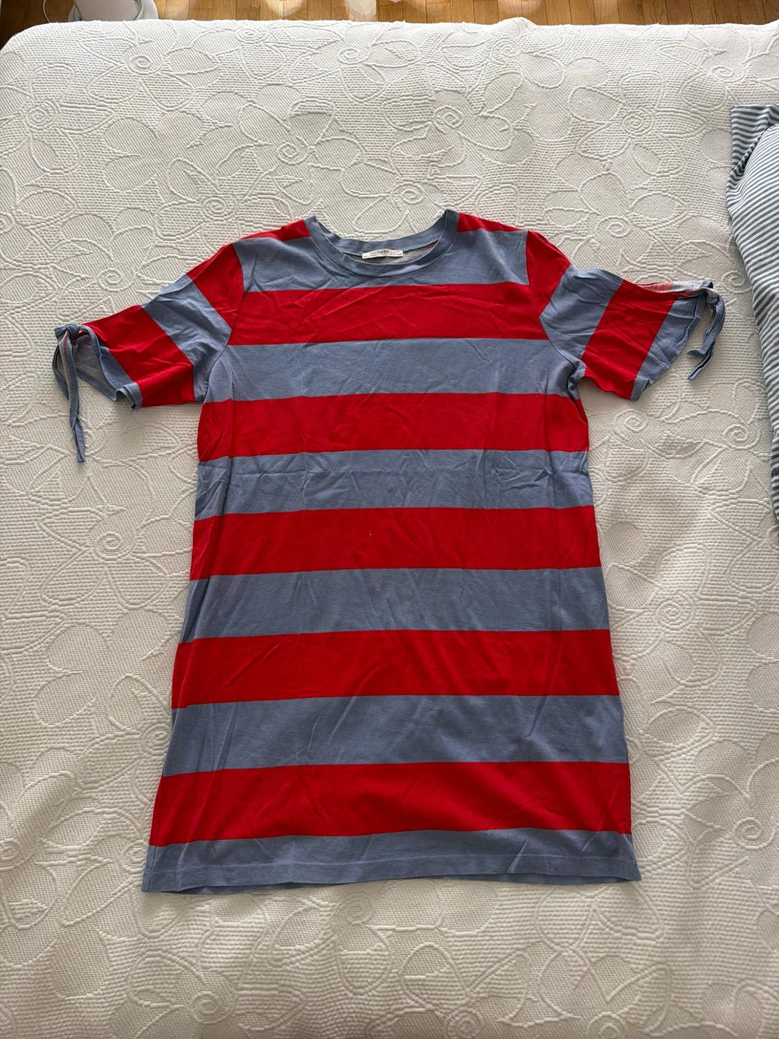 Red and blue horizontal striped t-shirt with bows on the sleeves, laid out on a textured white bedspread. Relaxed style.