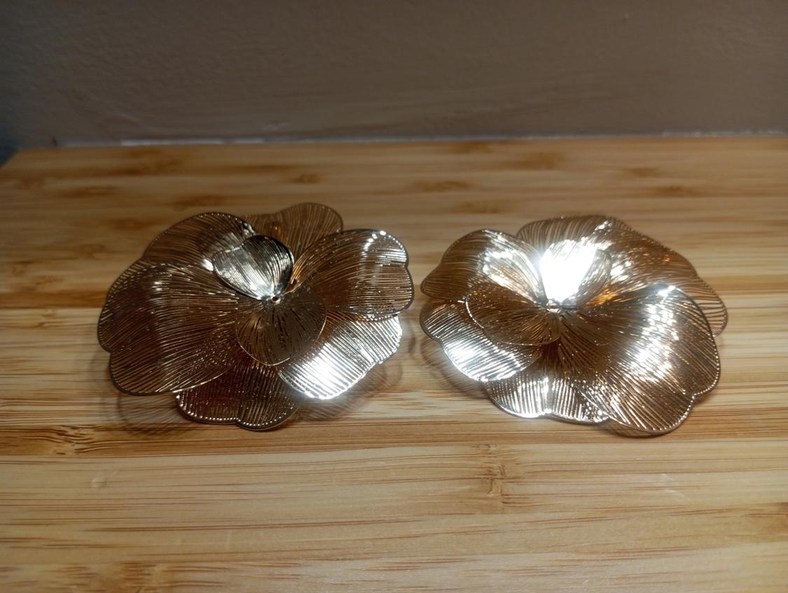 Gold flower-shaped earrings on a light wooden surface, reflecting the light and showing textured details.