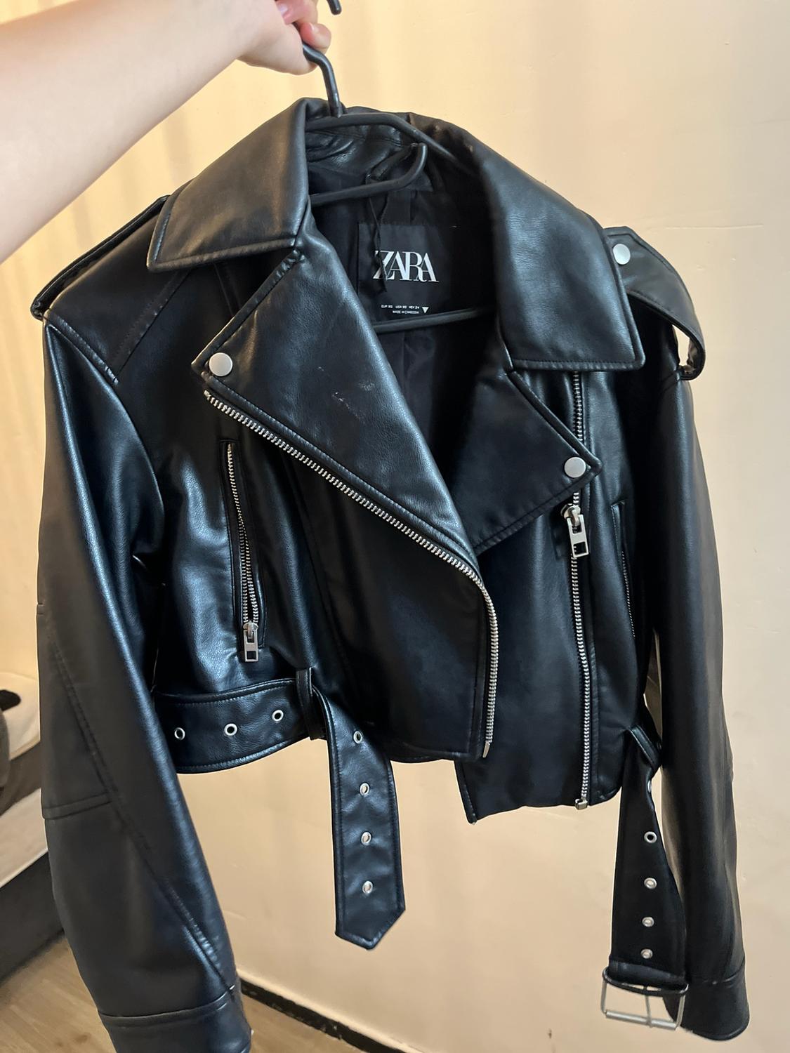 A hand holds a black jacket with silver zips and a belt hanging from a hanger. The label reads ZARA.