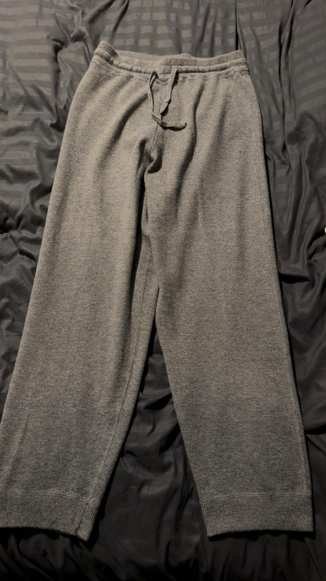 Grey knitted trousers with an elasticated waistband and adjustable drawstring, laid out on a creased dark fabric background.