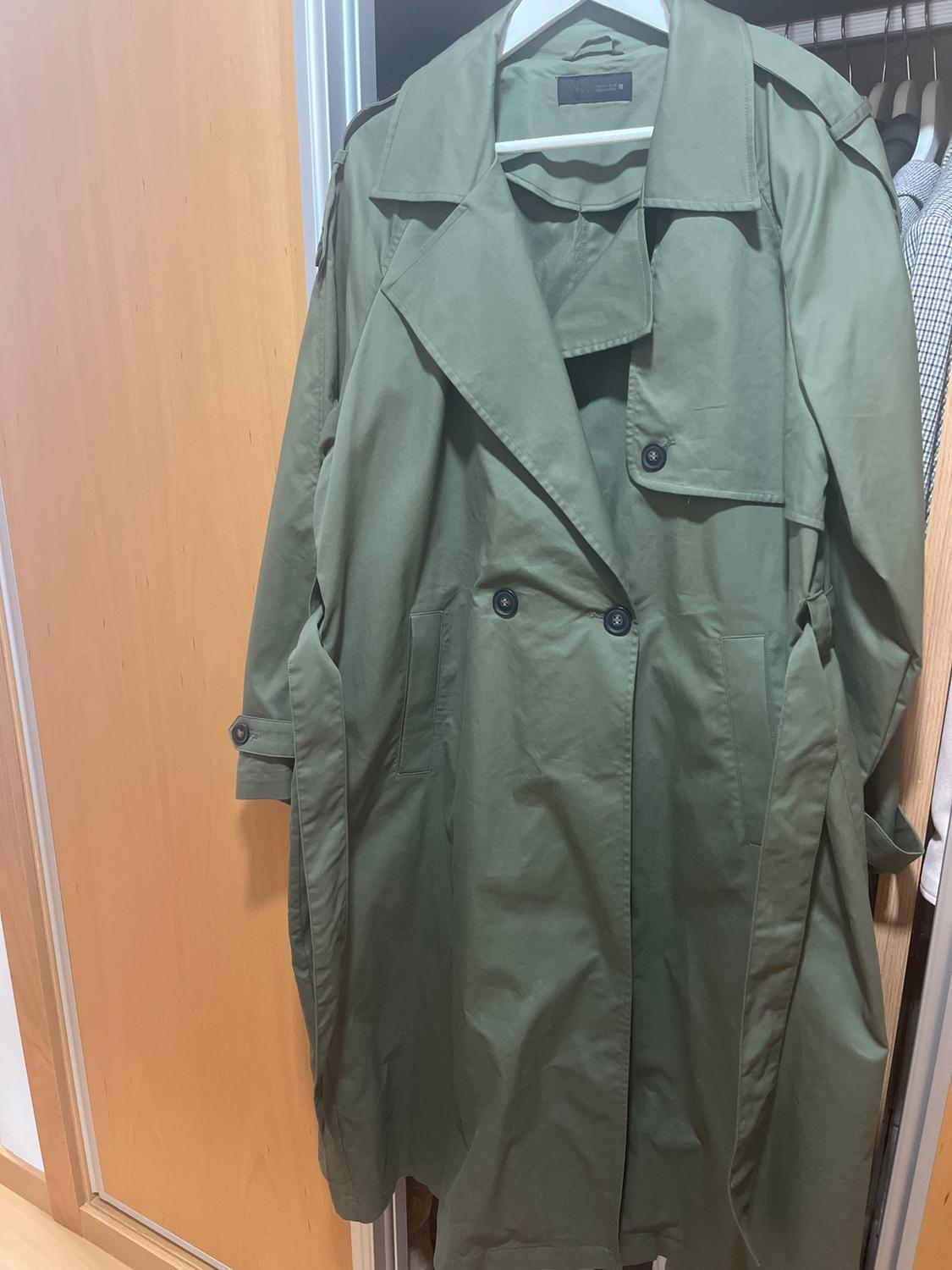 Olive green trench coat hanging in a wooden wardrobe, with black buttons and wide lapels. Background of hanging clothes.